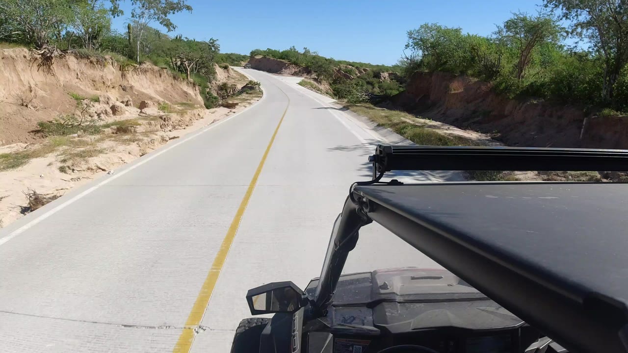 Forward view from a vehicle on a pale paved road in Los Cabos, Mexico, with sandy roadside cuts, green desert brush, blue sky, and no visible people.