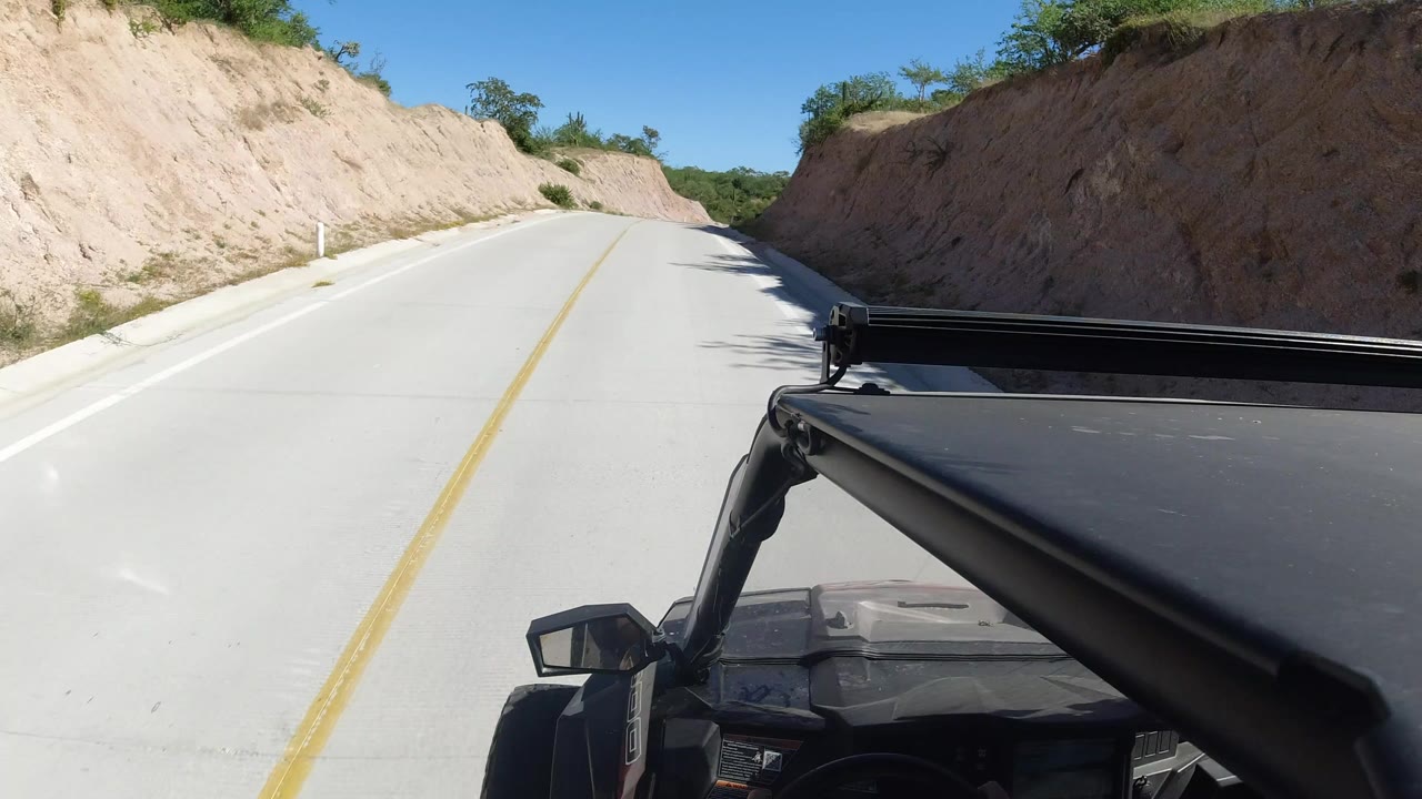 Opening frame of a Los Cabos road seen from a vehicle, with tan embankments, blue sky, a black roof edge in the foreground, and no visible people.
