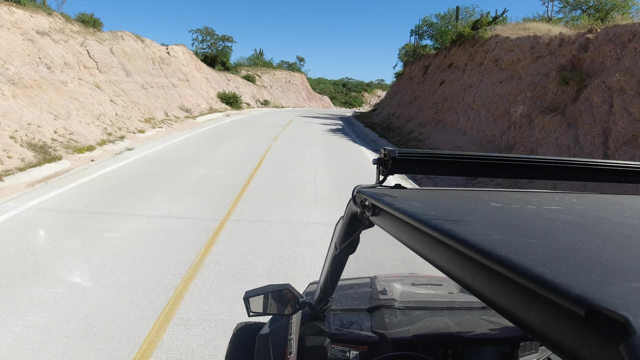 Road view from a vehicle passing a sandy embankment under clear sky.