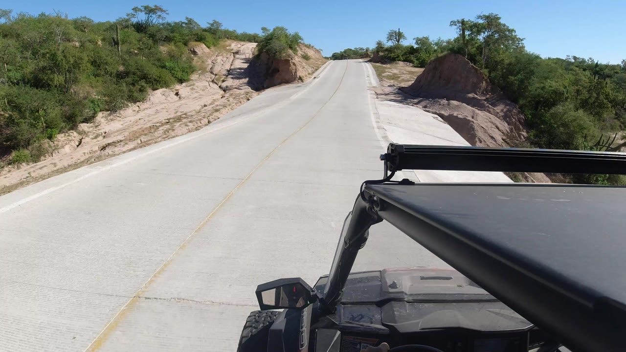 Forward view from a moving vehicle on a pale road in Los Cabos, Mexico, with sandy banks and green roadside vegetation, no visible people.