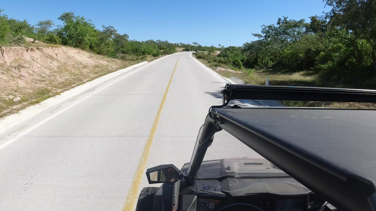 The Los Cabos road continues ahead under clear blue sky, with dense green vegetation on both sides and the SUV roof rack crossing the lower right foreground.