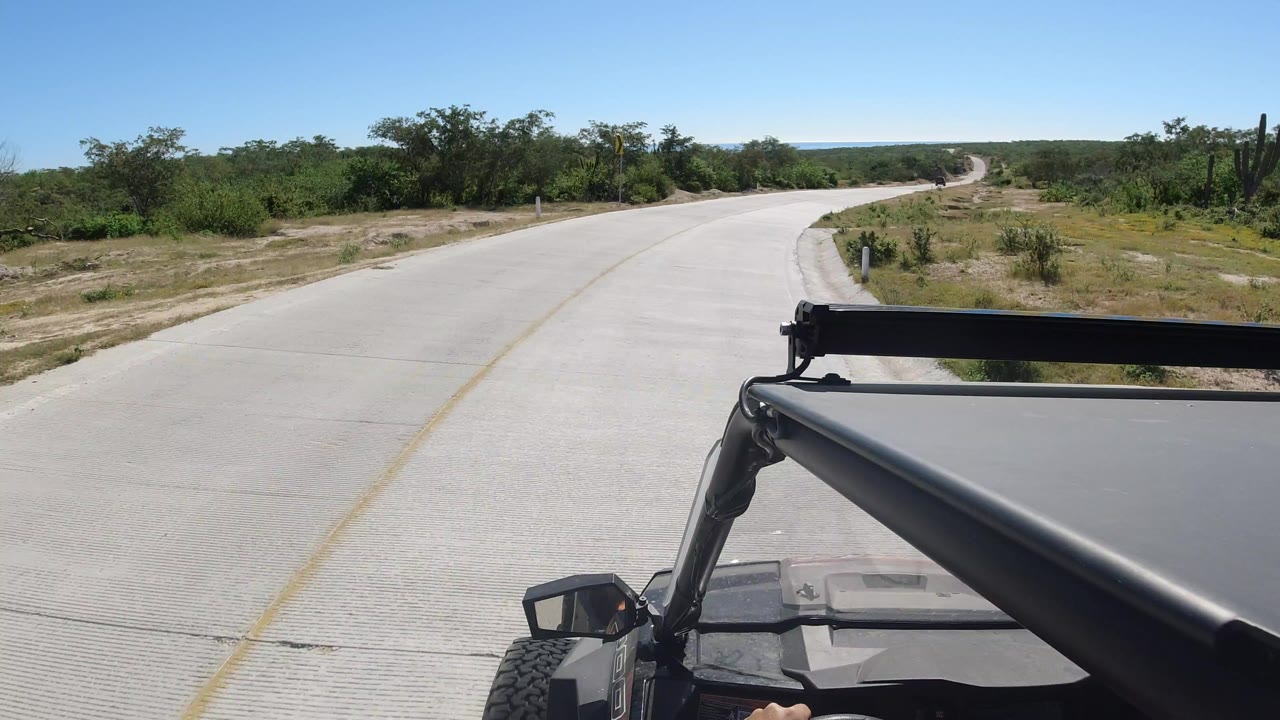 Forward view from a black vehicle on a paved road in Los Cabos, Mexico, with low green brush, cactus shapes, blue sky, and no visible people.