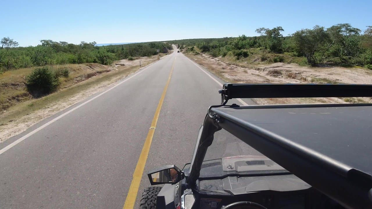Start of the Los Cabos, Mexico road clip, showing a straight paved road, yellow center line, vehicle roof hardware in the foreground, dry shoulders, green roadside trees, and no visible people.