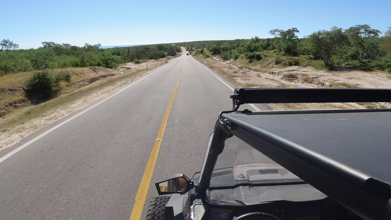 One second into the Los Cabos, Mexico clip, the road remains centered while the black roll bar and roof edge occupy the foreground and scrubby green vegetation lines both shoulders with no visible people.