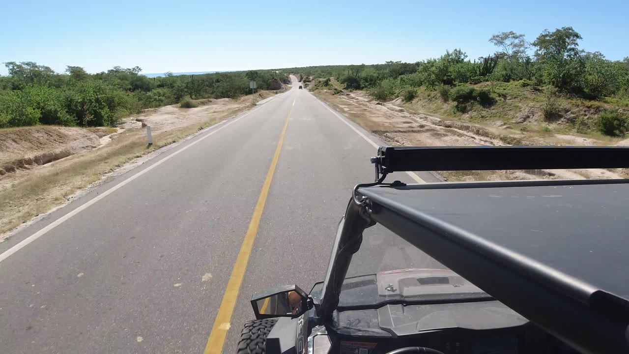 Empty road ahead with vehicle roof in foreground and scrub vegetation around it.