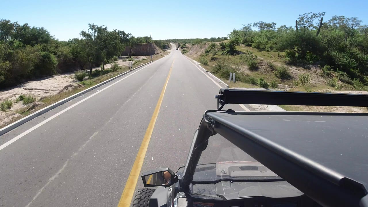 Paved rural road seen from a moving vehicle with scrub on both sides.