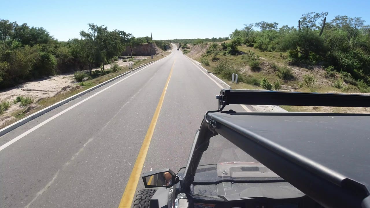 Forward vehicle view along a quiet Los Cabos road, with scrub vegetation on both shoulders, sandy ground beyond the pavement, and no visible people.