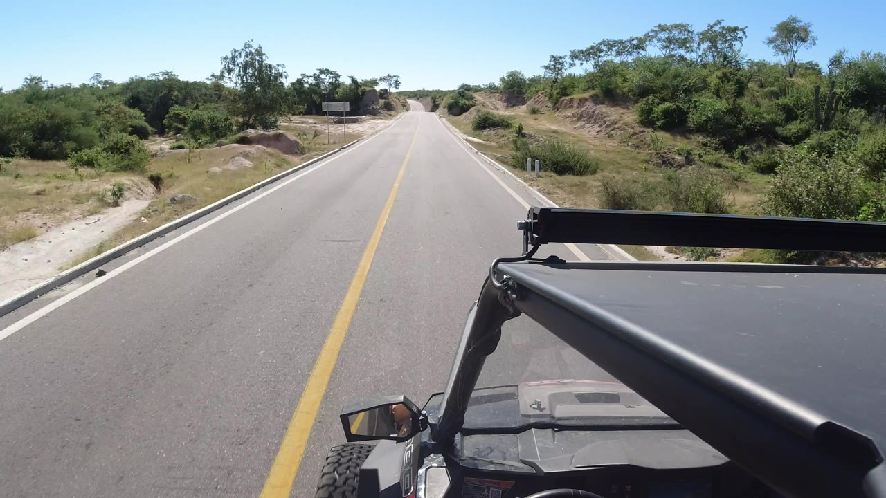 SUV-mounted view along a straight Los Cabos highway, with the road narrowing toward the horizon, scrub vegetation on both sides, and no visible people.