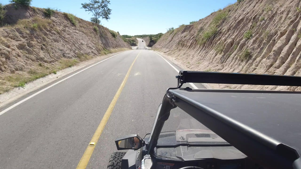 Forward view from a vehicle on a paved road between dry slopes.