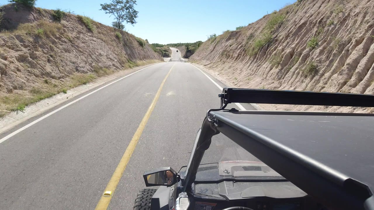 The road continues ahead in Los Cabos, Mexico, framed by eroded sandy banks, low roadside vegetation, the vehicle's roof edge and black bar in the foreground, and no visible people.