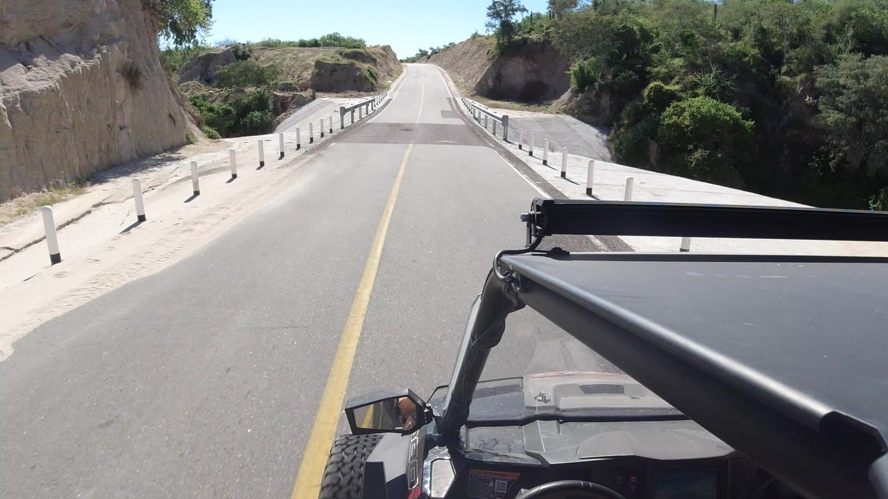 Forward vehicle POV in Los Cabos, Mexico, looking down a paved road toward a bridge with white roadside posts, dry cut hills, green brush, and no visible people.