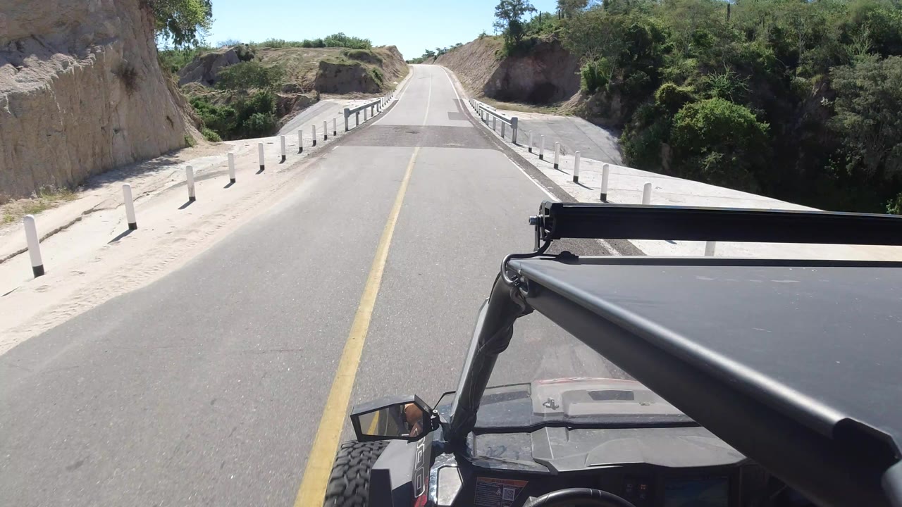 Vehicle-mounted view in Los Cabos, Mexico, centered on a yellow-lined road crossing a short bridge, with the vehicle hood low in frame and no visible people.