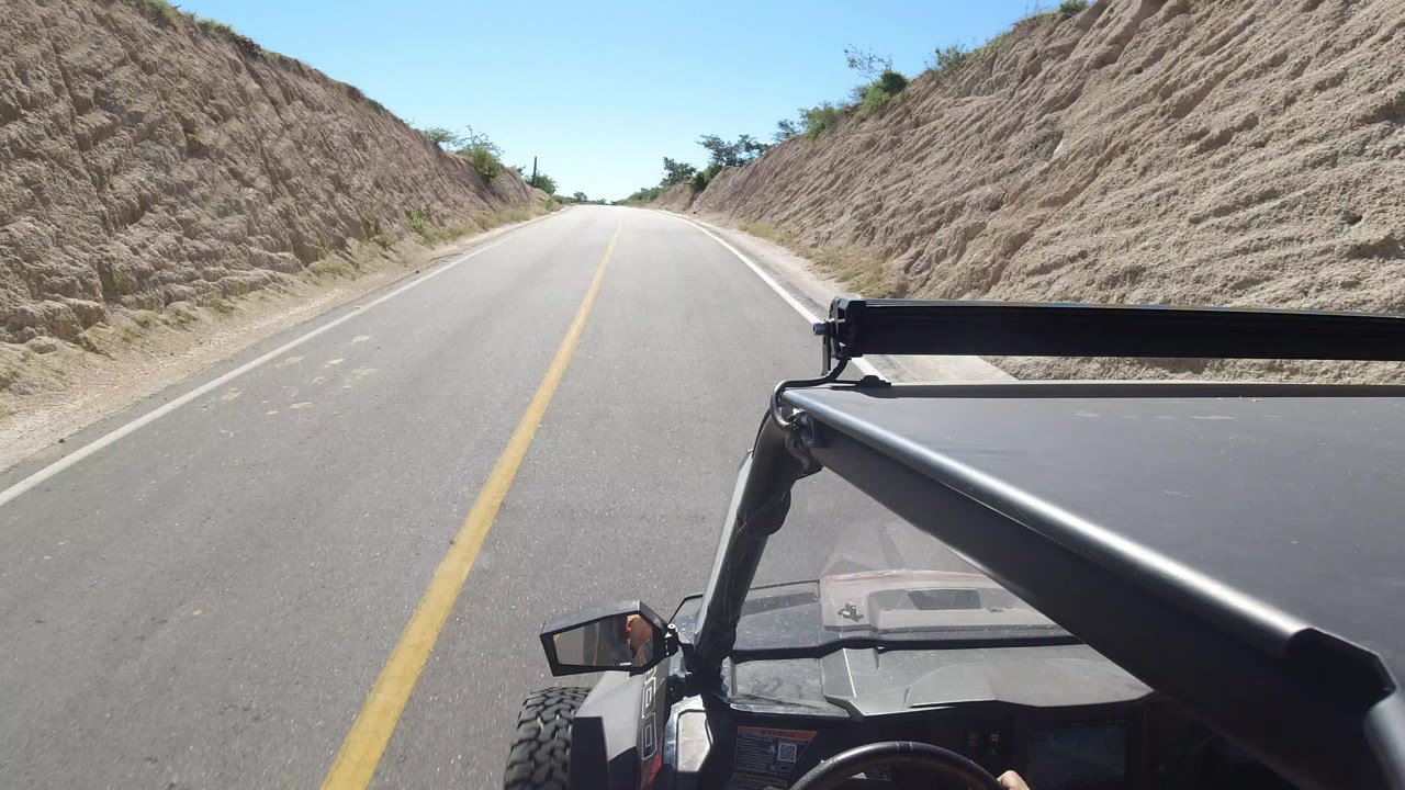 Opening view from a moving vehicle on a paved Los Cabos road, with sandy cut banks on both sides, blue sky overhead, and no visible people.