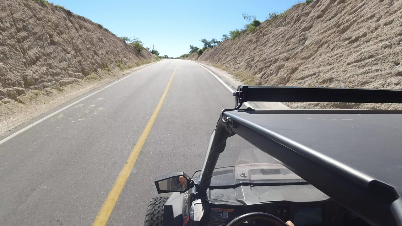 A later Los Cabos road POV shows the lane continuing between eroded sandy banks, with the open vehicle roll bar filling the lower right and no visible people.