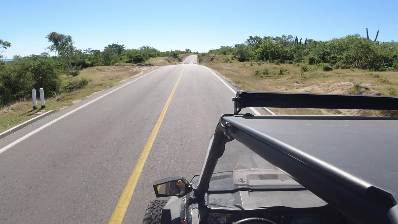 Forward vehicle-mounted view on a paved Los Cabos, Mexico road with a yellow center line, low green trees, dry shoulders, and no visible people.