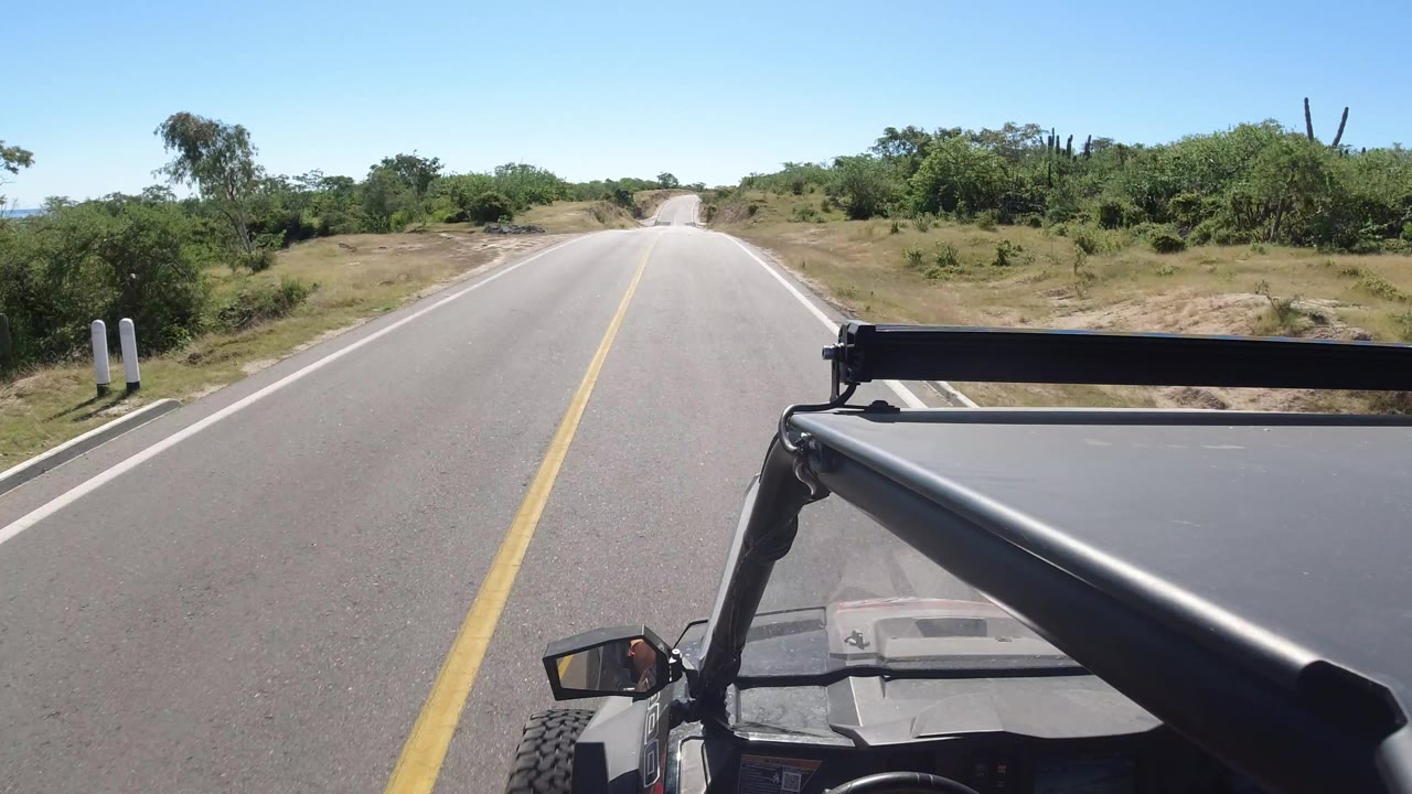 A centered road view in Los Cabos, Mexico shows the vehicle roof and roll bar in the foreground, scrubby trees on both sides, and no visible people.