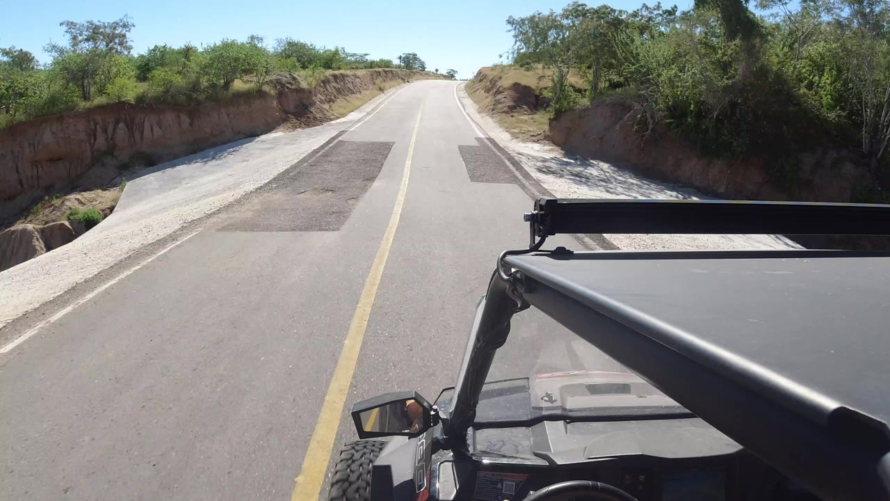 Forward view from an open vehicle on a paved road in Los Cabos, Mexico, with the hood in the foreground, a dirt bank at right, shrubs at left, and no clearly visible face.