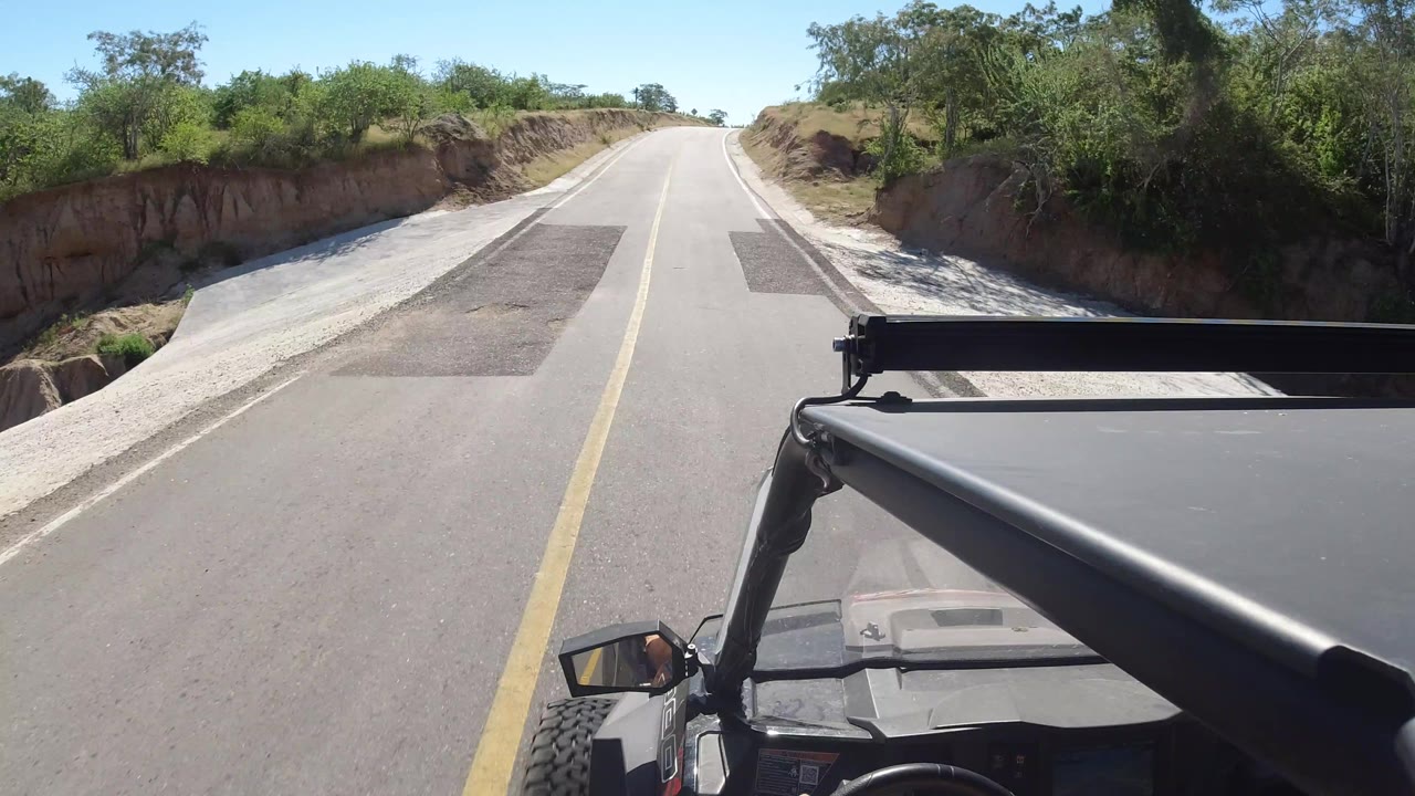 The vehicle continues along a two-lane road in Los Cabos, Mexico, with patched asphalt, scrub vegetation, and the front frame of the vehicle visible at the bottom.
