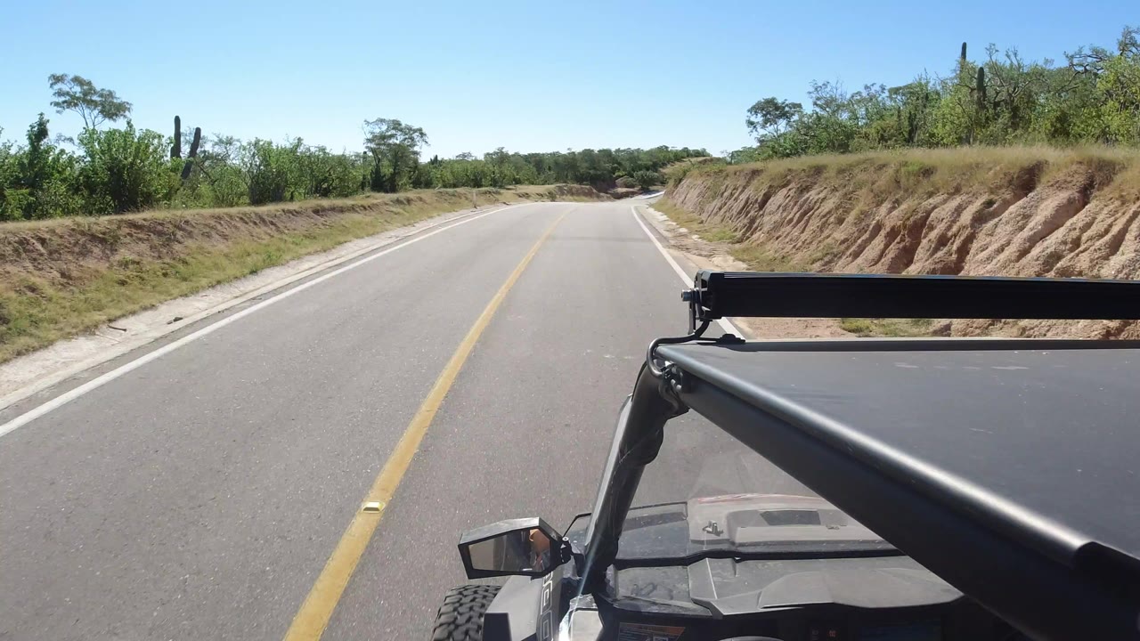 Vehicle-mounted view down a paved road with dry banks and green brush.