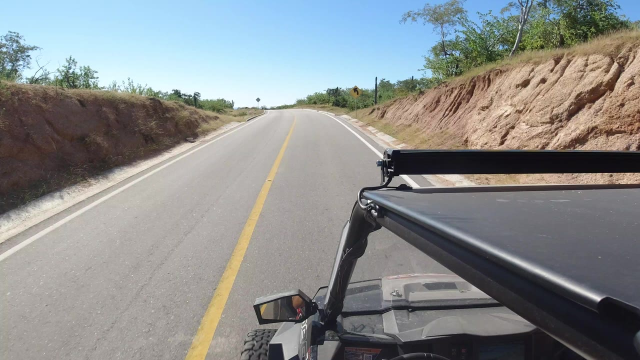 Forward road view in Los Cabos, Mexico with a paved lane between cut dirt banks, bright blue sky, vehicle roof rack in the foreground, and no visible people.
