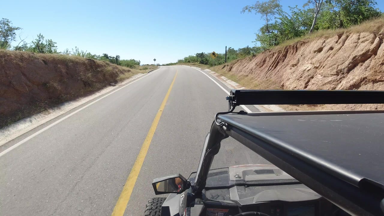 Forward-facing Los Cabos road frame with dry slopes on both sides, sparse trees above the cut banks, the vehicle corner at lower right, and no visible people.