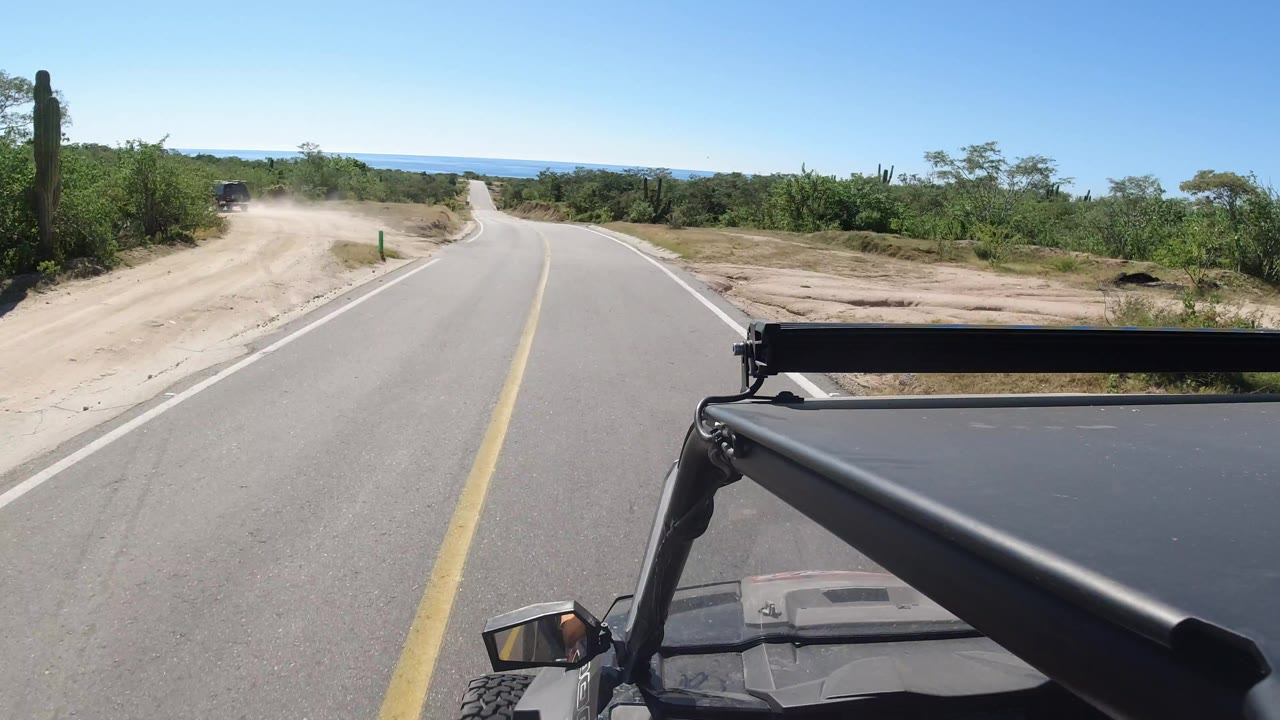 One second into the Los Cabos, Mexico drive, the forward view shows the road curving right, a dusty side road on the left, green brush, tall cactus, bright blue sky, and no visible people.