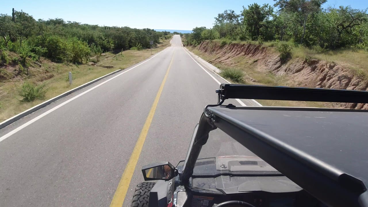 The SUV continues along a straight paved road in Los Cabos, Mexico, with low green vegetation, exposed roadside dirt banks, foreground vehicle parts, and no visible people.