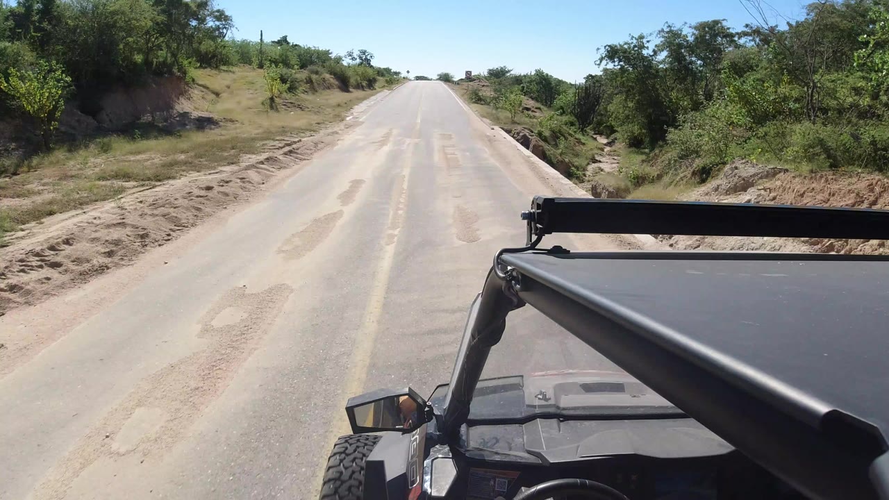 Forward vehicle POV on an asphalt road in Los Cabos, Mexico, with a yellow center line, roadside brush, bright sky, and no visible faces.