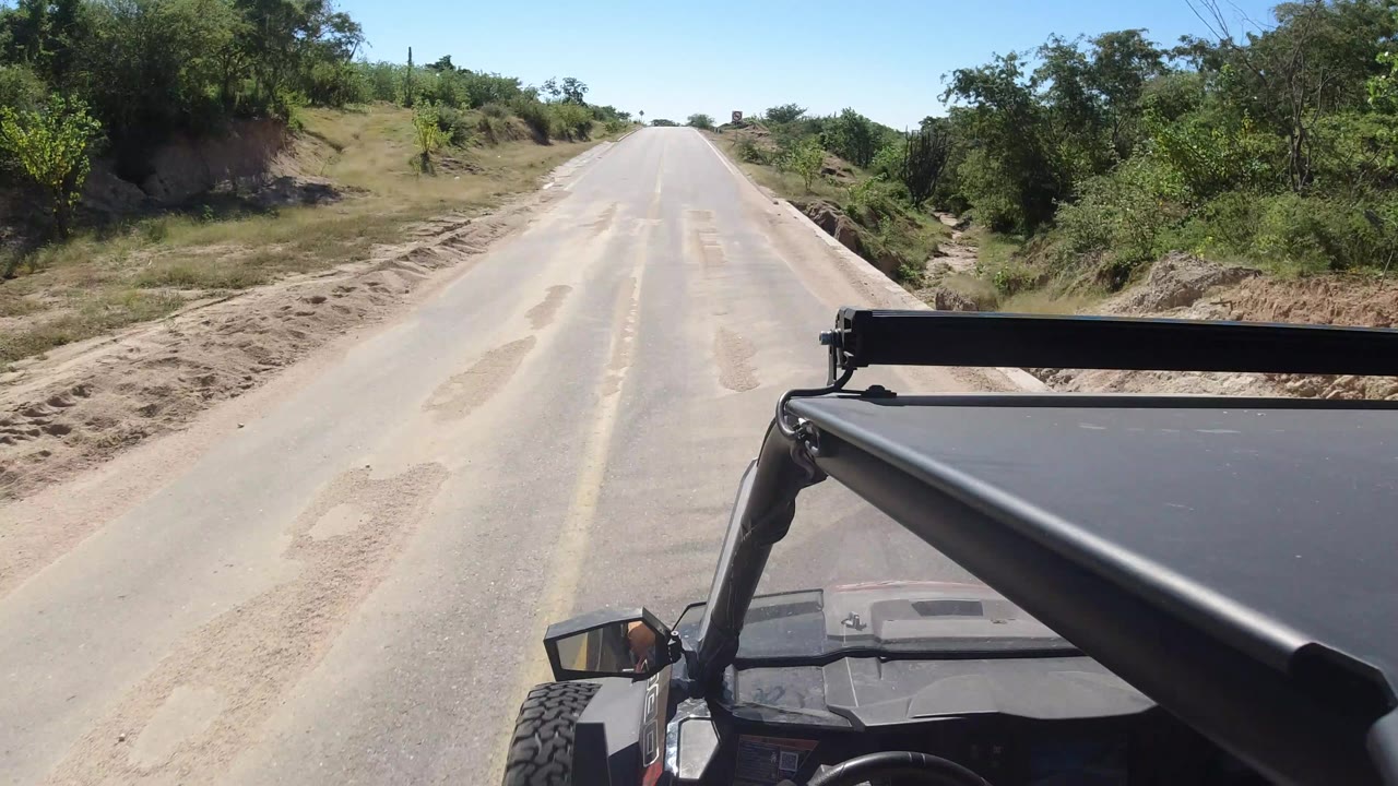 The road remains centered ahead in Los Cabos, Mexico, with scrub vegetation, exposed dirt shoulders, and the vehicle mirror and roof hardware visible at the lower right.