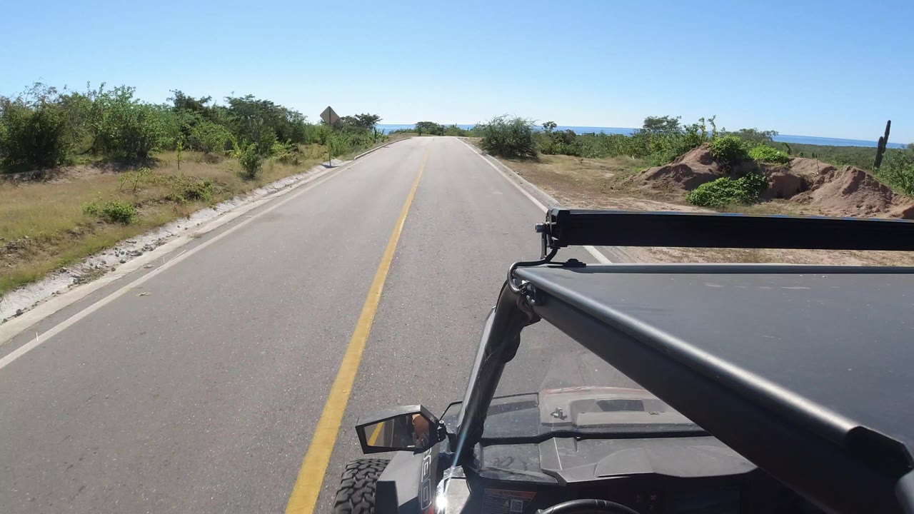 Forward view from a vehicle on a paved road with roadside bushes and distant water.