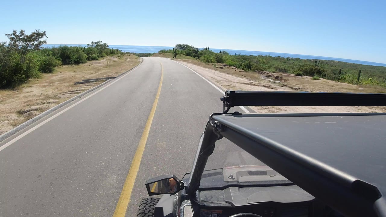 Forward view from a vehicle on a sunlit road in Los Cabos, Mexico, with the ocean ahead, dry roadside brush, and no visible people.