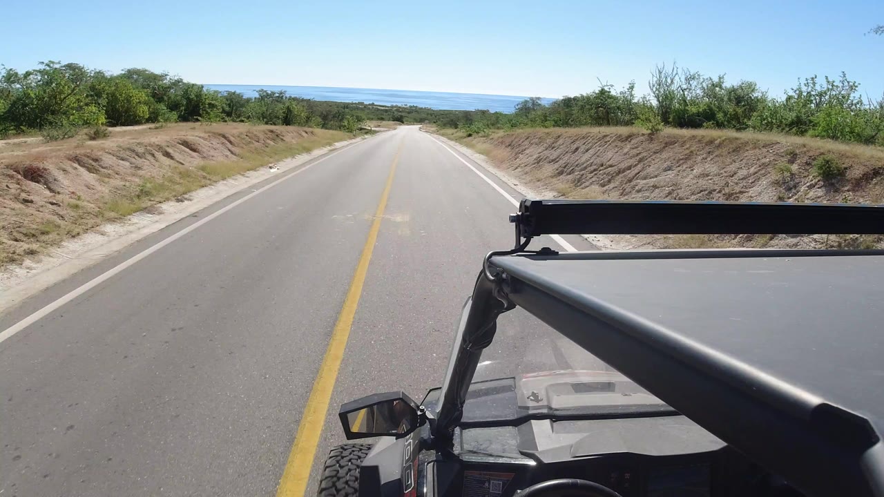 Start of the Los Cabos road clip, looking down a straight paved road from a vehicle roof area toward distant blue water, with low green brush on both sides and no visible people.