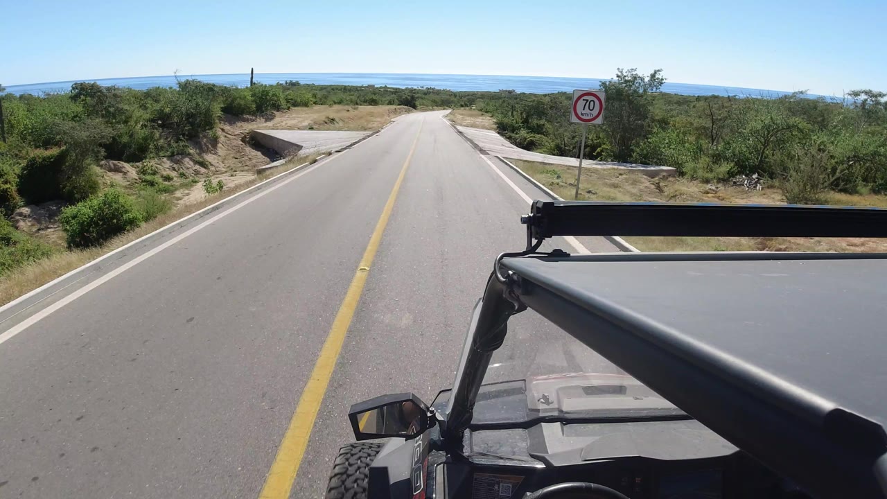 The vehicle continues along a Los Cabos highway, showing a straight stretch of pavement, a roadside 70 sign, scrub vegetation, distant water, and no visible people.