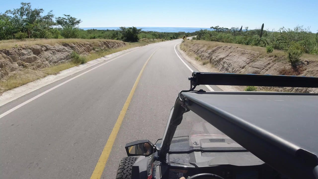 Opening view of a Los Cabos road curving ahead, with the vehicle hood and roll bar at lower right, dry roadside brush, distant water on the horizon, and no visible people.
