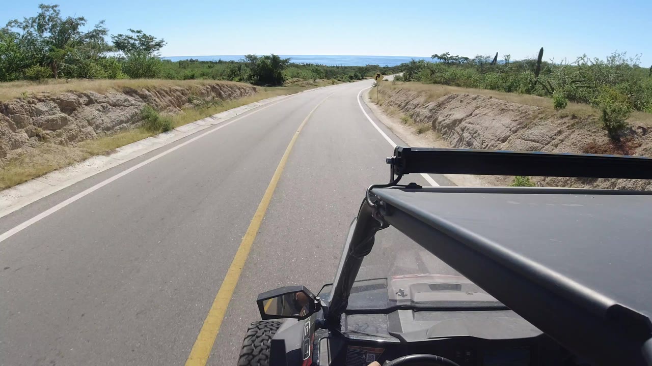 A forward road view in Los Cabos shows the yellow centerline leading downhill toward distant water, with the open vehicle frame at the lower right and no visible people.