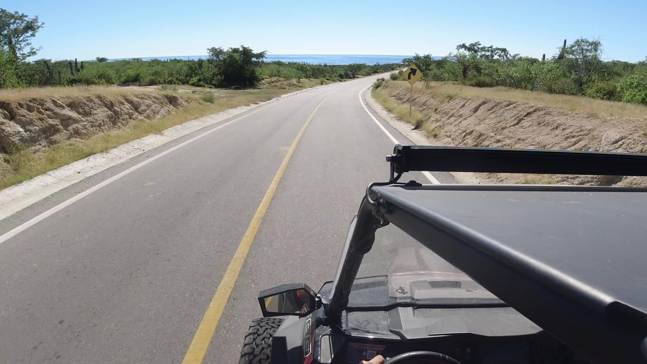 Road curve bordered by scrub vegetation and a visible vehicle hood.