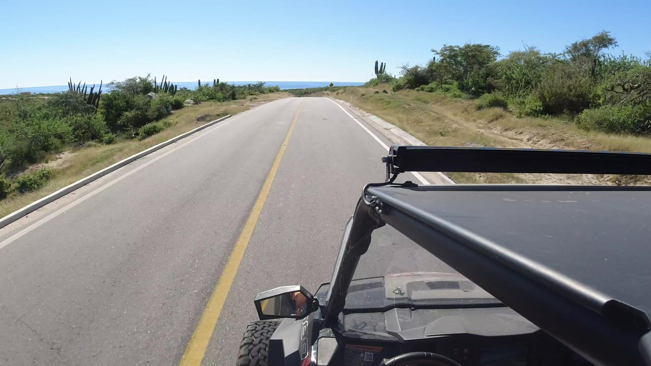 Forward-facing vehicle view on a paved Los Cabos road, with a yellow centerline, scrub vegetation, distant blue water, a black roll bar in the foreground, and no visible people.