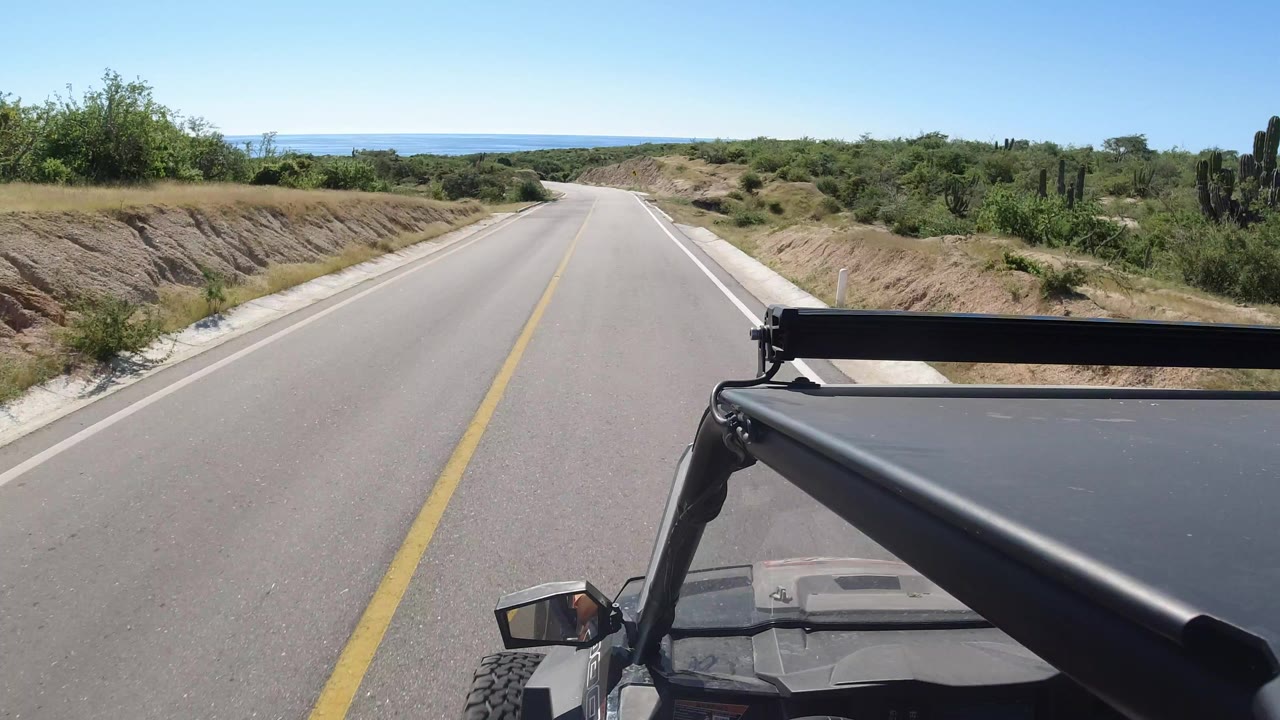 Front vehicle view on an empty Los Cabos road, with a black roof bar and windshield edge in the foreground, low green brush along both sides, distant water on the horizon, and no visible people.