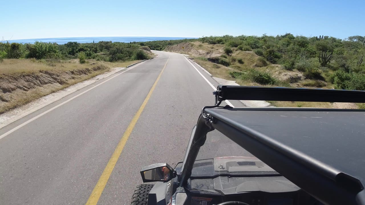 Empty road beginning to curve right with brush, sandy slopes, and vehicle bar in foreground.