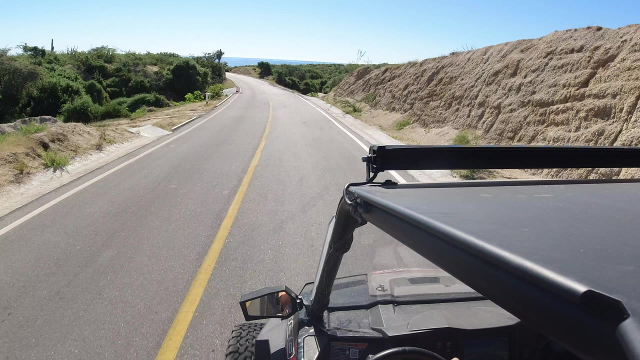 Forward vehicle view on a Los Cabos road with a yellow centerline, scrub vegetation, sandy bank, blue horizon, and no visible people.