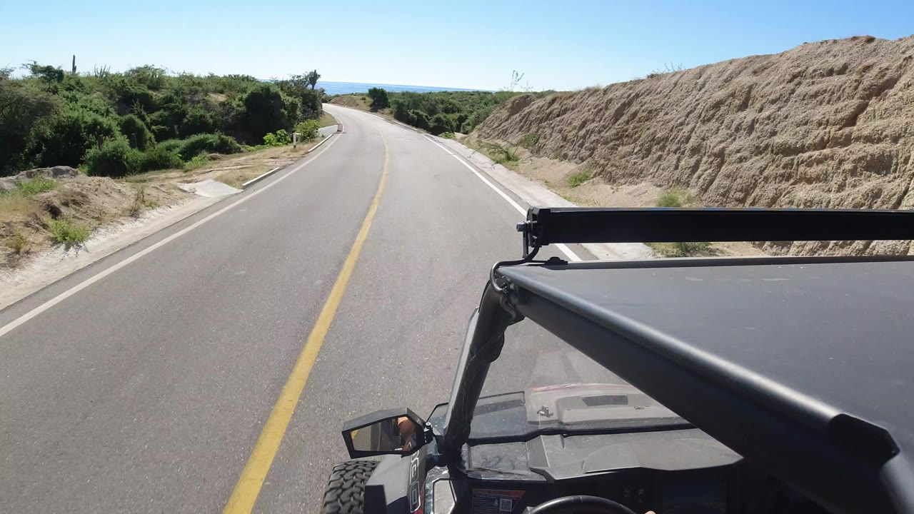 Los Cabos road view from an open vehicle, with the pavement curving left between dense green shrubs, a sandy slope, and no visible people.
