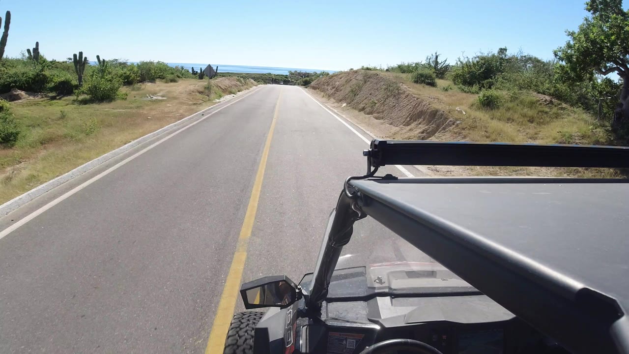 The jeep-mounted camera looks straight down the Los Cabos road as cactus and low green vegetation line both sides under bright blue sky, with no visible people.