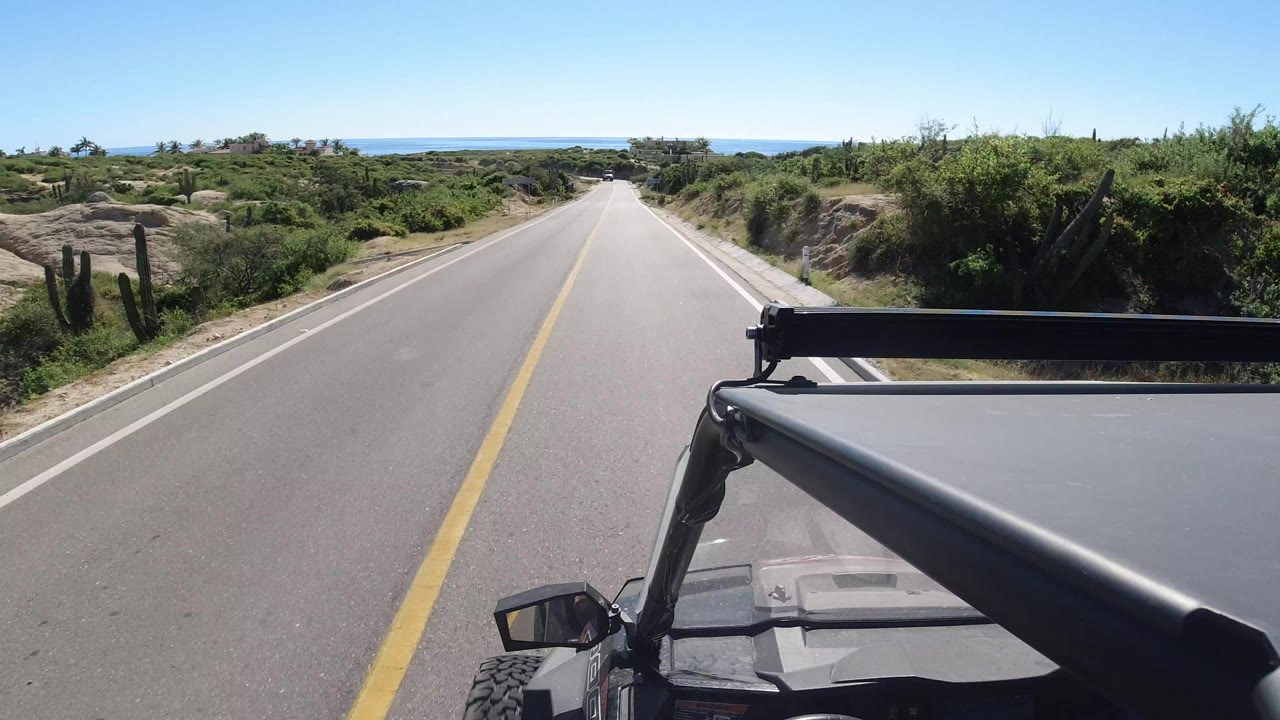 Start of a Los Cabos, Mexico road view over a black vehicle hood and roll bar, with a yellow centerline, low green brush, cactus, and a blue water horizon in the distance, no visible people.