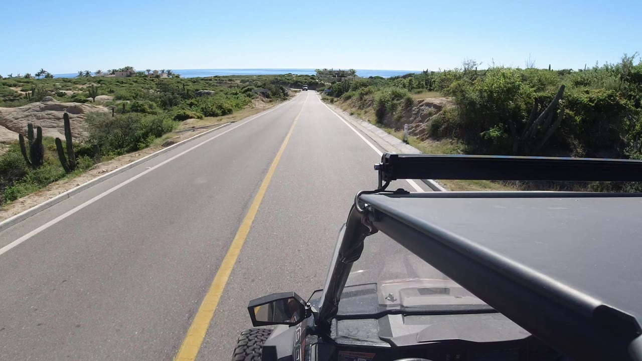 A centered Los Cabos road view continues past shrubs and sandy shoulders, with the black vehicle hood and roll bar steady in the foreground and the blue coastline visible ahead, no visible people.