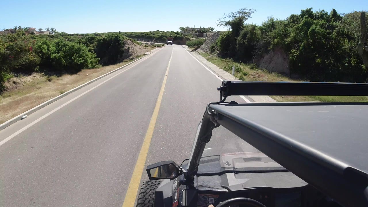Forward vehicle view along a sunlit road in Los Cabos, Mexico, with the open vehicle frame at lower right, roadside vegetation, sandy banks, blue sky, and no visible people.