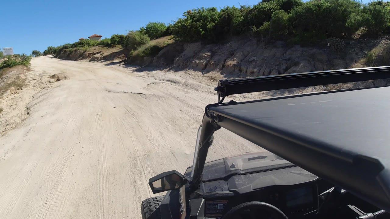 The Los Cabos road continues ahead between scrub-covered banks, with the vehicle roof and roll bar occupying the lower right and no visible people.