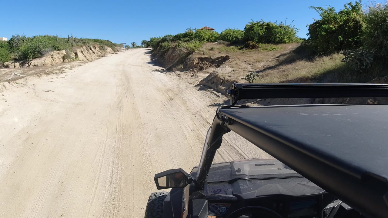 A Los Cabos roadside view shows the sandy track centered ahead, low dirt banks on both sides, thick green shrubs to the right, vehicle hardware in the foreground, and no visible people.