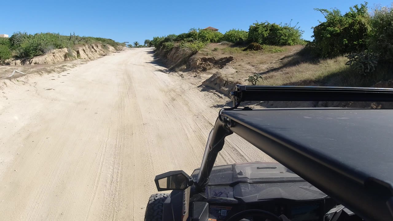 Sandy dirt road ahead of an open vehicle with green bushes on both sides.