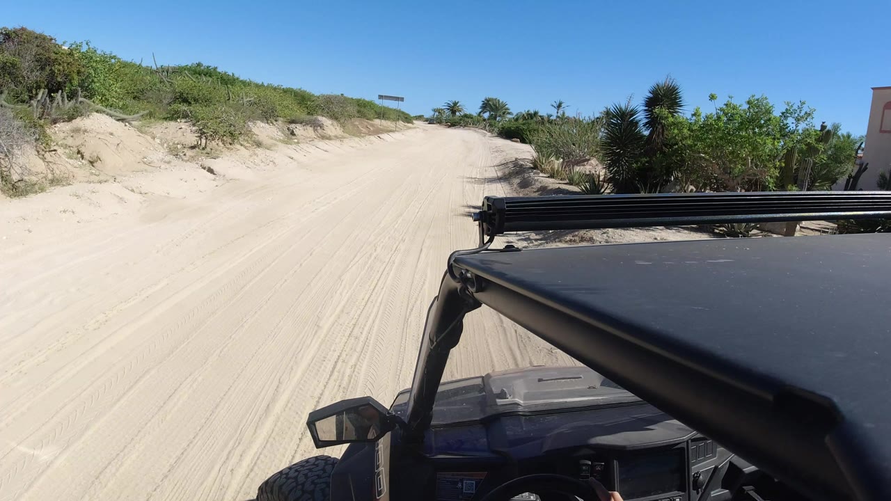 A first view from a buggy in Los Cabos, Mexico shows the black roof and light bar at right, pale tire-marked sand ahead, low dunes, green brush, and no visible people.
