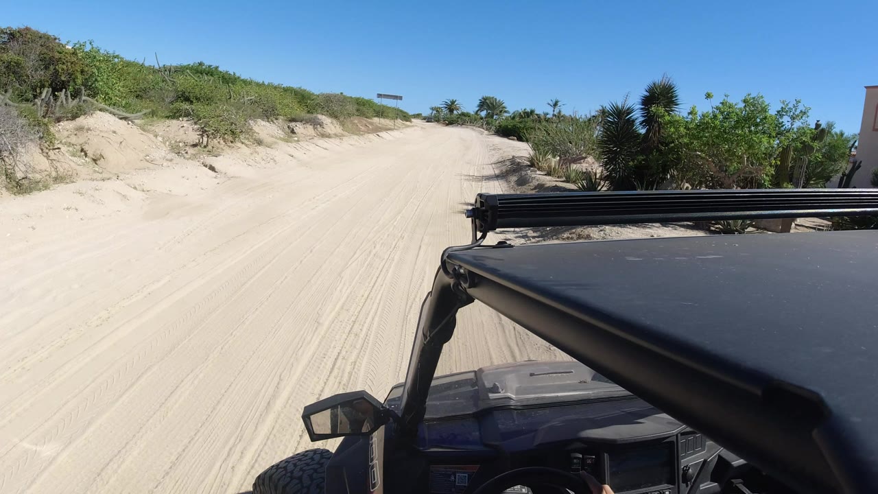 The buggy foreground stays at right while the sandy Los Cabos track opens ahead between low dune banks, scrub vegetation, blue sky, and no visible people.